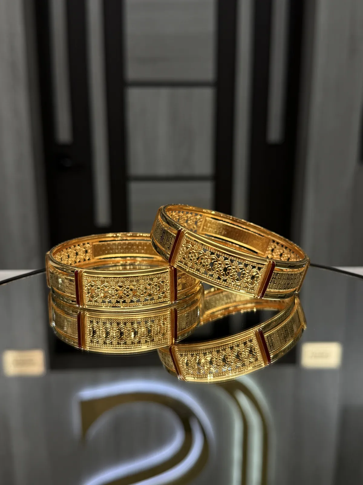 Pair of rectangular filigree 22K gold bangles with red accents on a mirrored display table